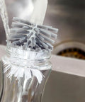 Clear bottle with a brush inside, likely used for cleaning purposes, against a blurred kitchen sink background.