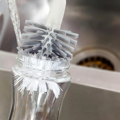 Clear bottle with a brush inside, likely used for cleaning purposes, against a blurred kitchen sink background.