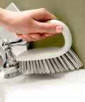 Hand holding a white scrub brush over a sink with a neutral background
