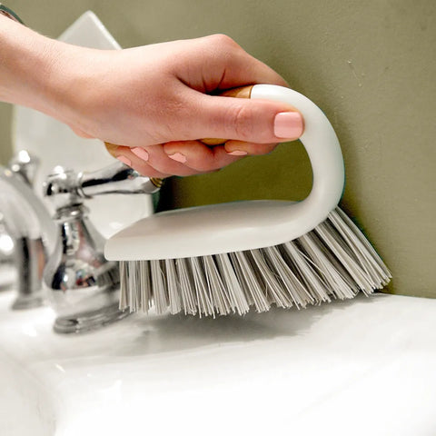 Hand holding a white scrub brush over a sink with a neutral background