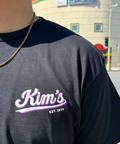 A close-up of a person wearing a dark charcoal gray t-shirt with a 'Kim's EST. 1979' graphic in white and purple on the chest.