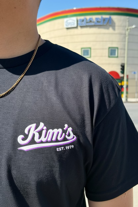 A close-up of a person wearing a dark charcoal gray t-shirt with a 'Kim's EST. 1979' graphic in white and purple on the chest.