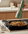 Person seasoning food in a pan with a green pepper grinder labeled 'finamill' in a kitchen setting.