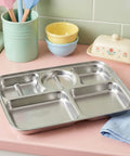 Stainless steel compartmentalized tray on a pink surface with colorful bowls and a green cup in the background.