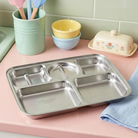 Stainless steel compartmentalized tray on a pink surface with colorful bowls and a green cup in the background.