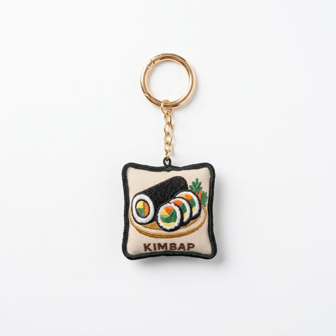 A square embroidered keychain charm featuring a depiction of kimbap, with a gold-tone split ring and chain, on a plain white background.
