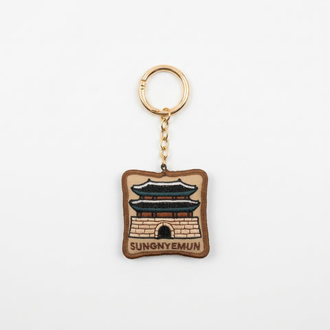 A square embroidered keychain charm featuring a brown and beige depiction of Sungnyemun gate, with a gold-tone split ring and chain, on a plain white background.