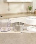 Kitchen countertop with a clear plastic container, metal strainer, and white lid on a beige surface.