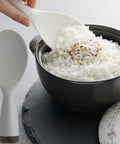 White rice being scooped into a black bowl with a white rice paddle on a gray surface.