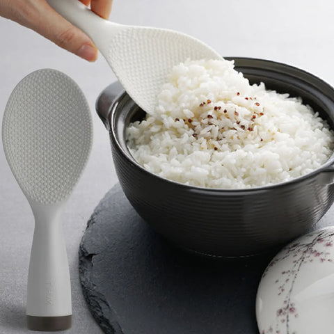 White rice being scooped into a black bowl with a white rice paddle on a gray surface.