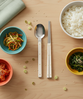 Dishes of food including rice, vegetables, and kimchi on a wooden table with utensils.