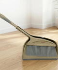 Beige broom and dustpan set on a wooden floor with a white wall and plant in the background.