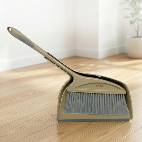 Beige broom and dustpan set on a wooden floor with a white wall and plant in the background.