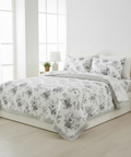 A neatly made bed in a bright, minimalist bedroom featuring a white and gray floral-patterned quilt, matching pillow shams, and a striped flat sheet.