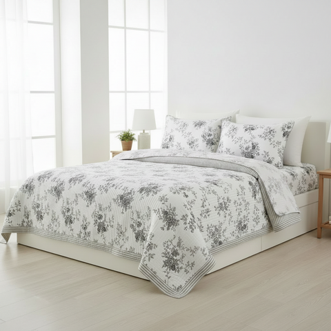 A neatly made bed in a bright, minimalist bedroom featuring a white and gray floral-patterned quilt, matching pillow shams, and a striped flat sheet.