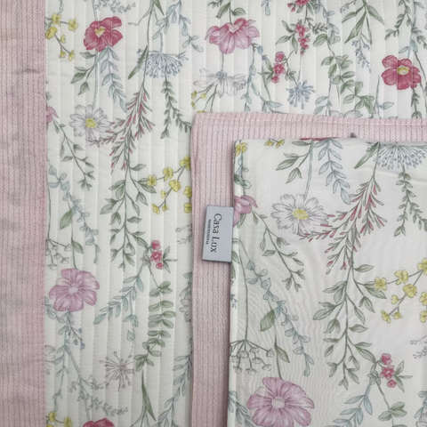 A close-up of a quilted fabric with a delicate pastel floral print on a cream background, featuring pink, green, and yellow flowers with a pink stitched border. A small fabric tag reading 'Casa Lux' is visible.