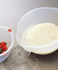 A clear plastic kitchen strainer bowl with a red handle is shown pouring rice into a sink while another bowl with vegetables sits beside it.