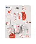 Anomata measuring cup in its original retail packaging, featuring a clear glass measuring cup with red measurement markings on a white card with red and black illustrations.