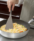 A person wearing an apron uses a gray silicone spatula with a textured surface to stir-fry rice with vegetables in a silver non-stick pan on a stovetop.