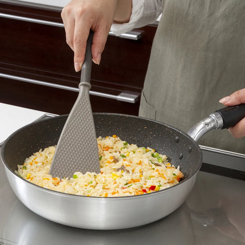 A person wearing an apron uses a gray silicone spatula with a textured surface to stir-fry rice with vegetables in a silver non-stick pan on a stovetop.