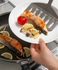 A hand holds a white plate with a cooked salmon slice, lemon wedges, mushrooms, and a small red tomato over a black frying pan on a stovetop. A gray silicone spatula rests on the plate.