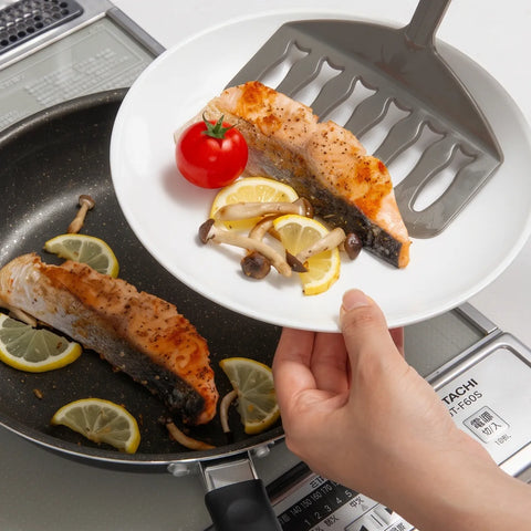 A hand holds a white plate with a cooked salmon slice, lemon wedges, mushrooms, and a small red tomato over a black frying pan on a stovetop. A gray silicone spatula rests on the plate.