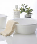 A white ceramic washbasin with a cream-colored textured washcloth draped over the side, set on a glossy white surface. In the softly blurred background, a clear bottle labeled BOTANIST and a small green plant are visible.