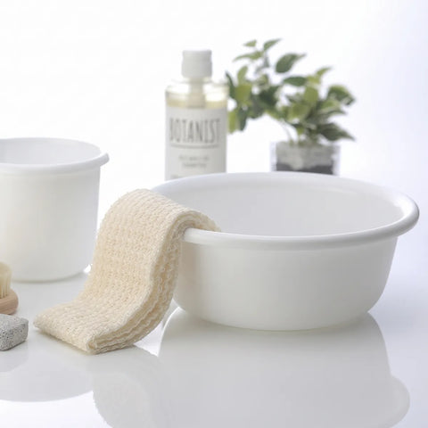 A white ceramic washbasin with a cream-colored textured washcloth draped over the side, set on a glossy white surface. In the softly blurred background, a clear bottle labeled BOTANIST and a small green plant are visible.