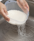 A clear plastic washbowl being held by two hands over a stainless steel sink, with white rice inside and water draining into the sink.