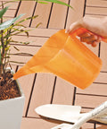 An orange translucent plastic watering can being held by a hand and used to water a potted plant on a brown wooden deck, with white garden trowels visible in the foreground.