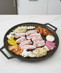 Round grill pan with sliced meat, vegetables, and other ingredients on a white background