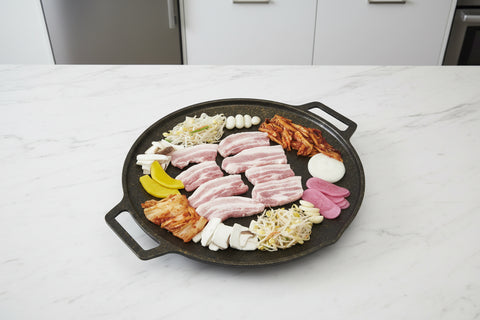Round grill pan with sliced meat, vegetables, and other ingredients on a white background