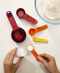 Colorful measuring spoons and cups on a white surface with hands using them.