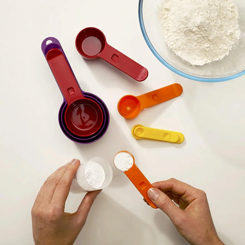 Colorful measuring spoons and cups on a white surface with hands using them.