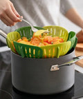 Person using a green kitchen steamer to transfer vegetables from a pot on a stove.