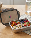 Metal lunch box with compartments containing food on a wooden table.