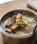 Steaming bowl of soup with noodles, vegetables, and meat in a stainless steel food container on a wooden table.