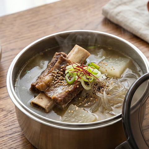 Steaming bowl of soup with noodles, vegetables, and meat in a stainless steel food container on a wooden table.