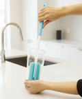 A person uses a blue and white cleaning brush to scrub inside a clear glass cup on a white kitchen countertop near a sink.