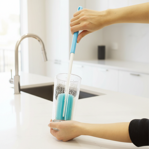 A person uses a blue and white cleaning brush to scrub inside a clear glass cup on a white kitchen countertop near a sink.