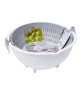 A white plastic spin-and-wheel colander basket with a gray interior, shown on a clean white background. Inside the basket are packaged food items, including a green pickle jar and a packet of Spin & Wheel Colander.