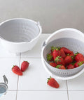 A white plastic strainer basket with a lid and side handles, shown empty and filled with red strawberries on a white tiled surface against a light gray wall.