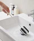 A hand holds a long-handled cleaning brush with a black and white striped sponge head inside a white bathroom sink, with soap dispensers and a faucet in the background.