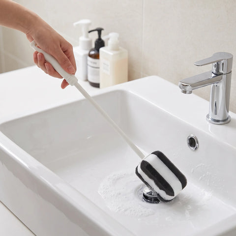 A hand holds a long-handled cleaning brush with a black and white striped sponge head inside a white bathroom sink, with soap dispensers and a faucet in the background.