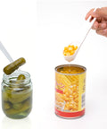 Two hands demonstrate a plastic spoon scoop against a white background, with one hand scooping yellow corn from a can and the other holding a pickle from a glass jar with the spoon.