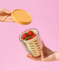 Hand holding a portable food container with strawberries and a hand lifting the yellow lid against a pink background