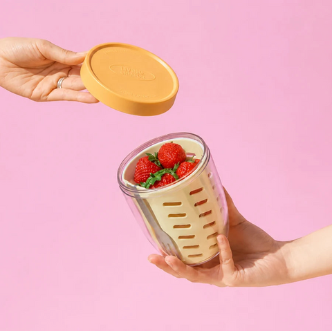 Hand holding a portable food container with strawberries and a hand lifting the yellow lid against a pink background