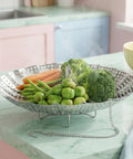 Vegetable steamer basket with carrots, broccoli, and green beans on a kitchen counter.