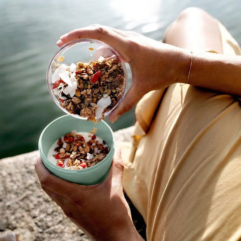 Person pouring granola into a bowl by a body of water