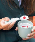 Person holding a small container of yogurt with a spoon, surrounded by red berries.